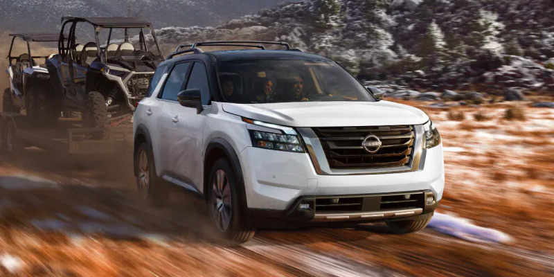 A white 2025 Nissan Pathfinder with a black roof confidently tows a trailer carrying two utility terrain vehicles on a dirt road through a mountainous, snowy landscape.