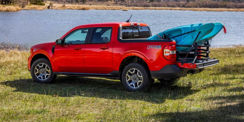 a red 2025 Ford Maverick truck exterior with a blue kayak in the bed parked in grass next to a lake